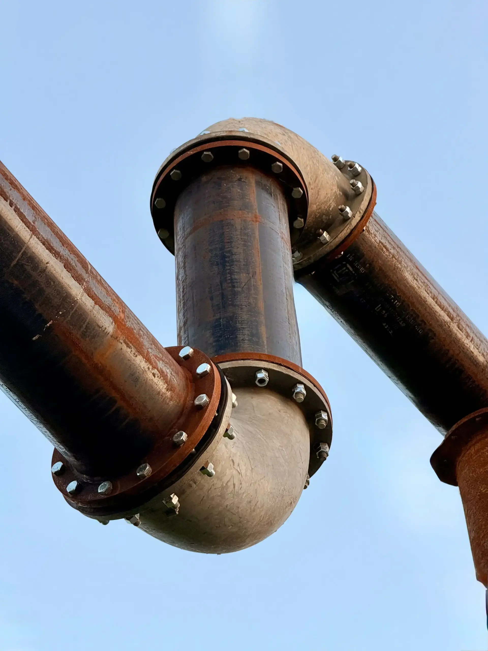 Industrial steel piping with bolted flange joints bending overhead against a blue sky